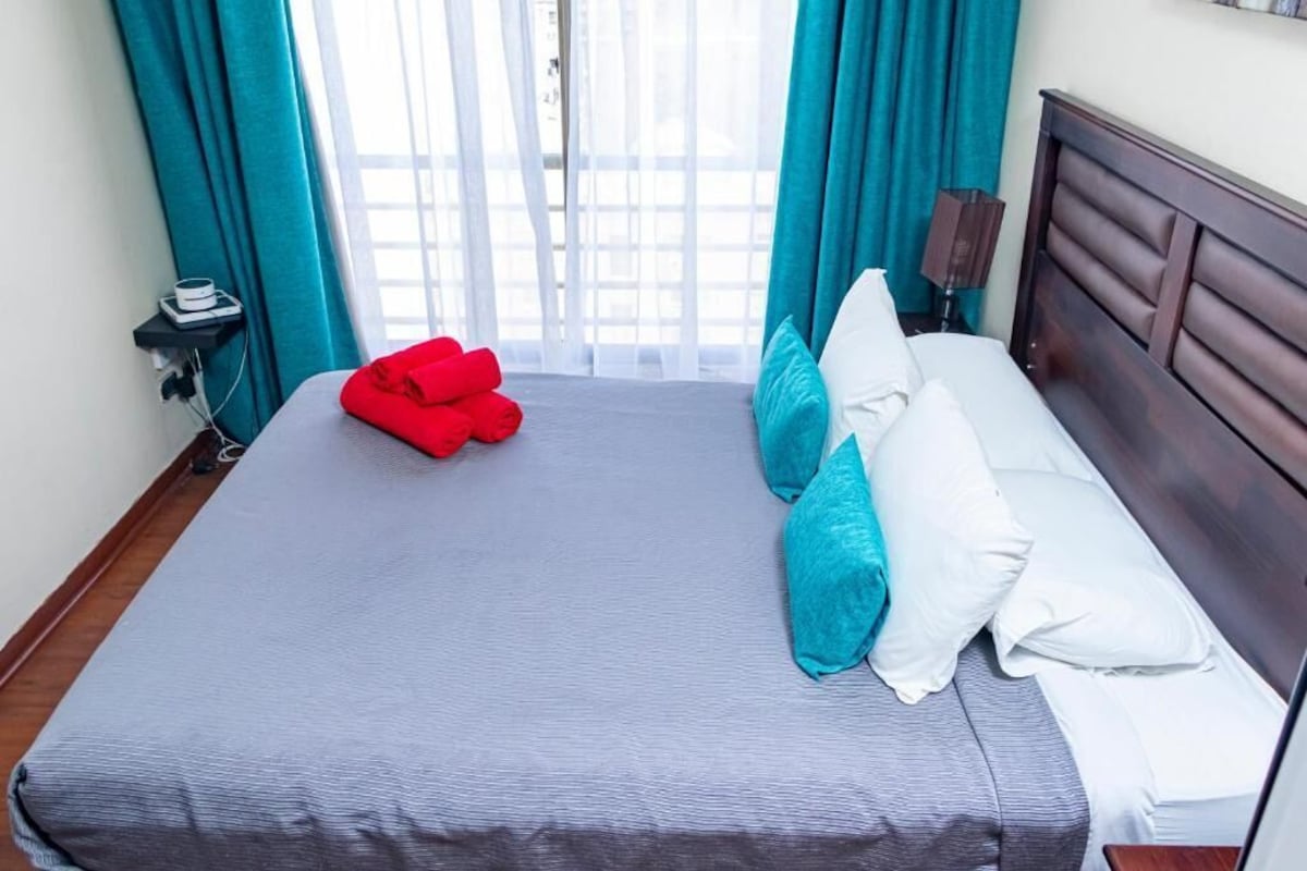 Un lit bien fait, recouvert d’une couverture grise, avec deux coussins turquoise et une serviette rouge.