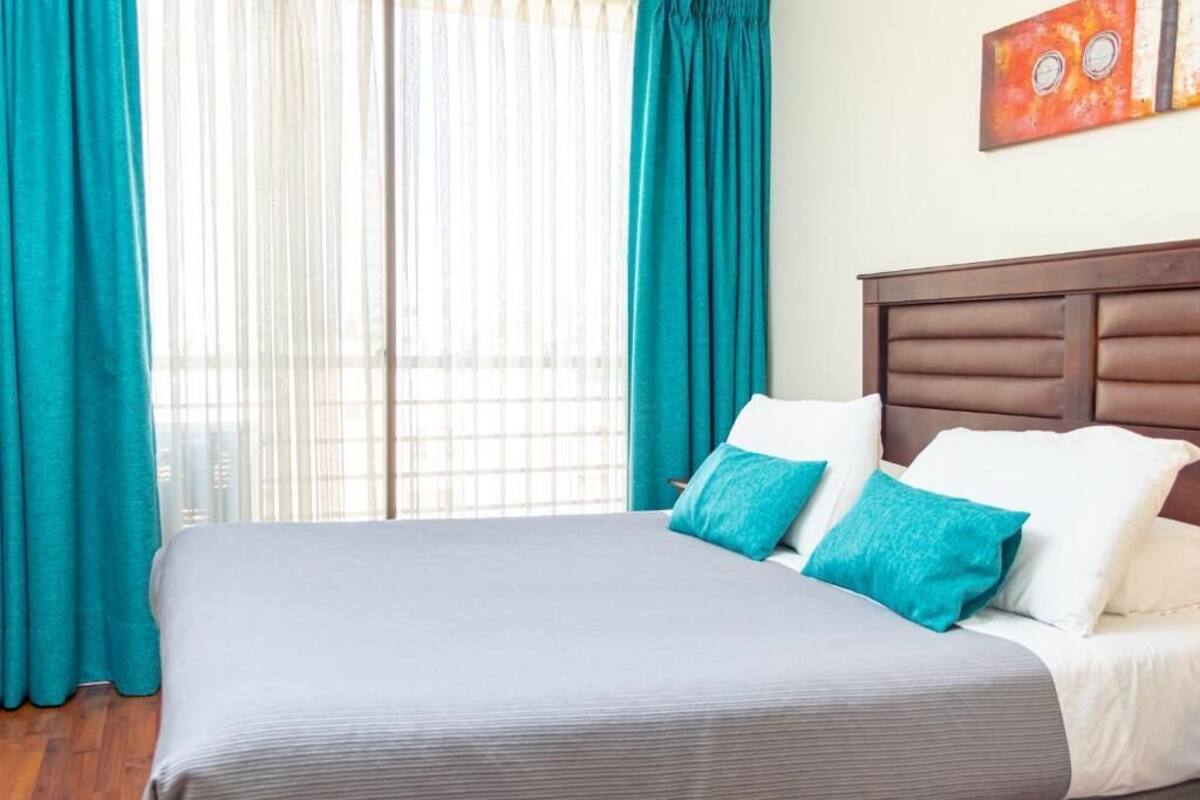 Habitació d'hotel amb un llit, cortines color aquamarina i una finestra amb cortines de tela fina.