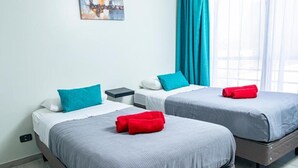 Een hotelkamer met twee bedden, elk voorzien van een grijze spreidek en teal kussens. Er is een nachtkastje met een lamp, een raam met teal gordijnen en een schilderij aan de muur.