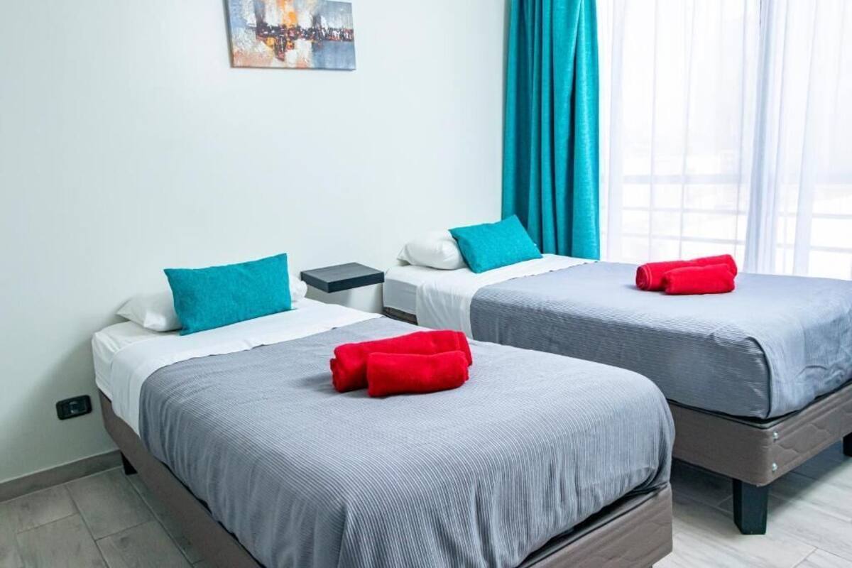 Habitació d'hotel amb dos llits, cadascun amb un llençol gris i almoades verda-blavosa. Hi ha una tauleta de nit amb una làmpada, una finestra amb cortines verda-blavoses i un quadre a la paret.