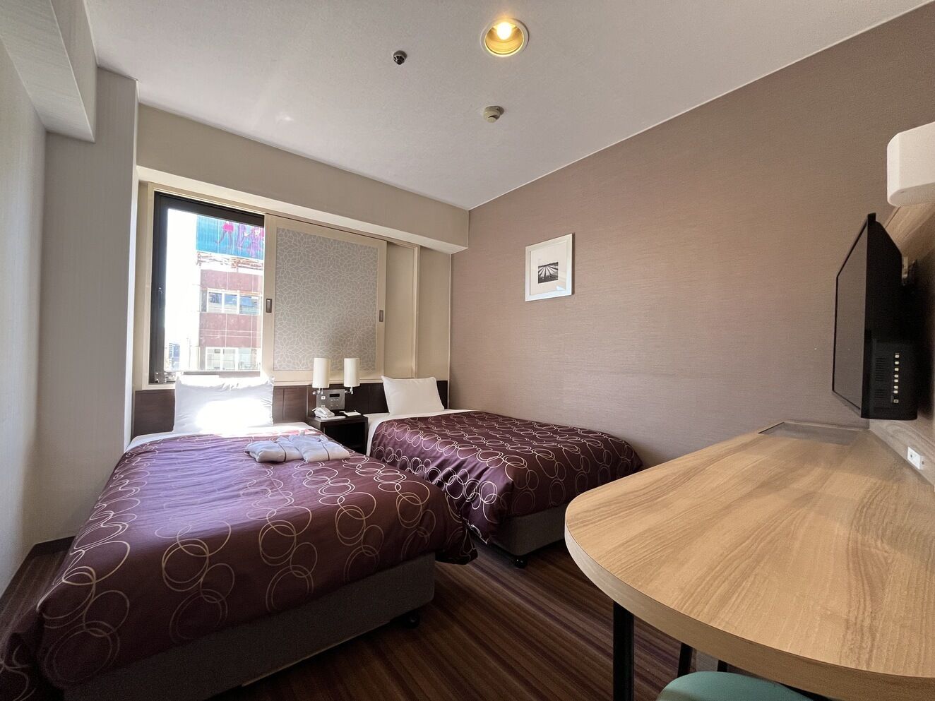 A hotel room with two beds, a small table, a TV, and a window with a view of buildings.