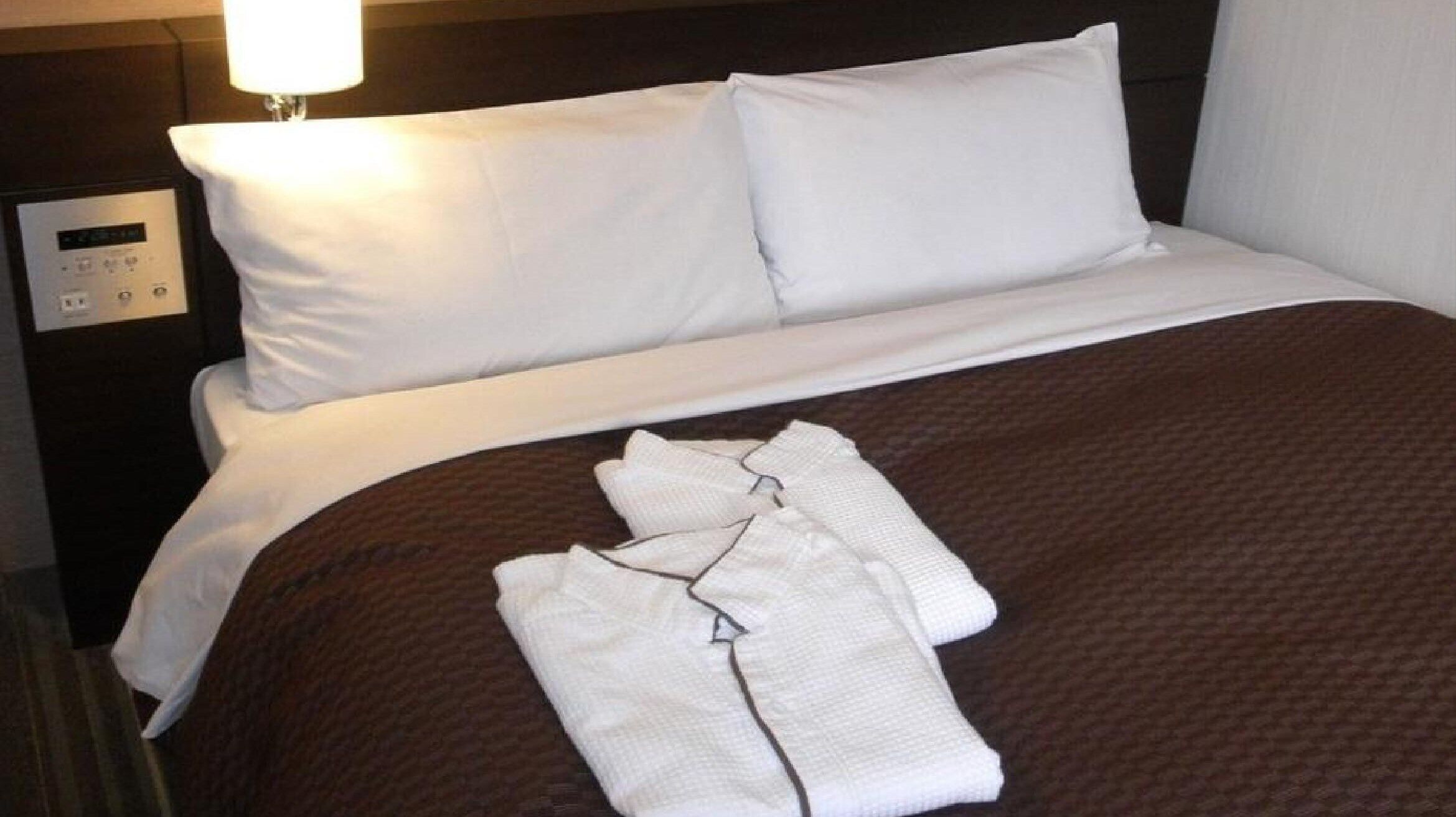 A neatly made bed with white linens, a brown blanket, and a folded white garment on the bed.
