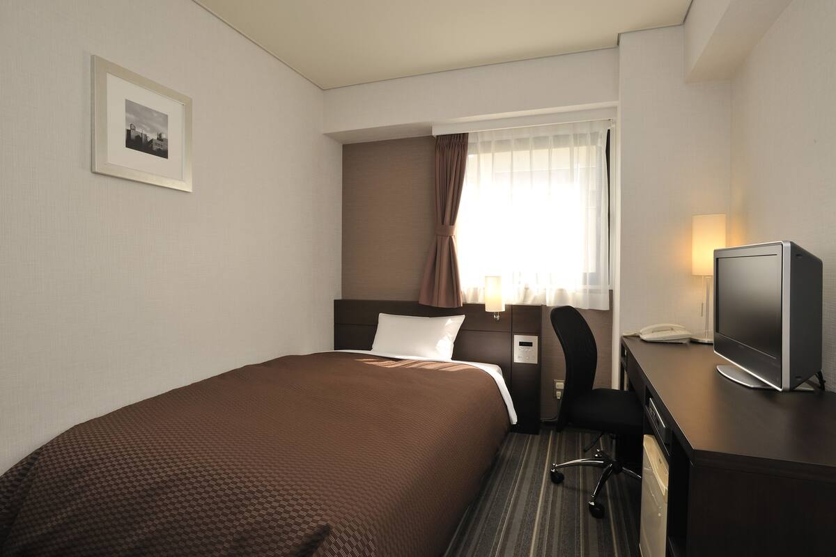 Quarto de hotel com cama, escrivaninha com computador, cadeira, abajur e janela com cortinas.