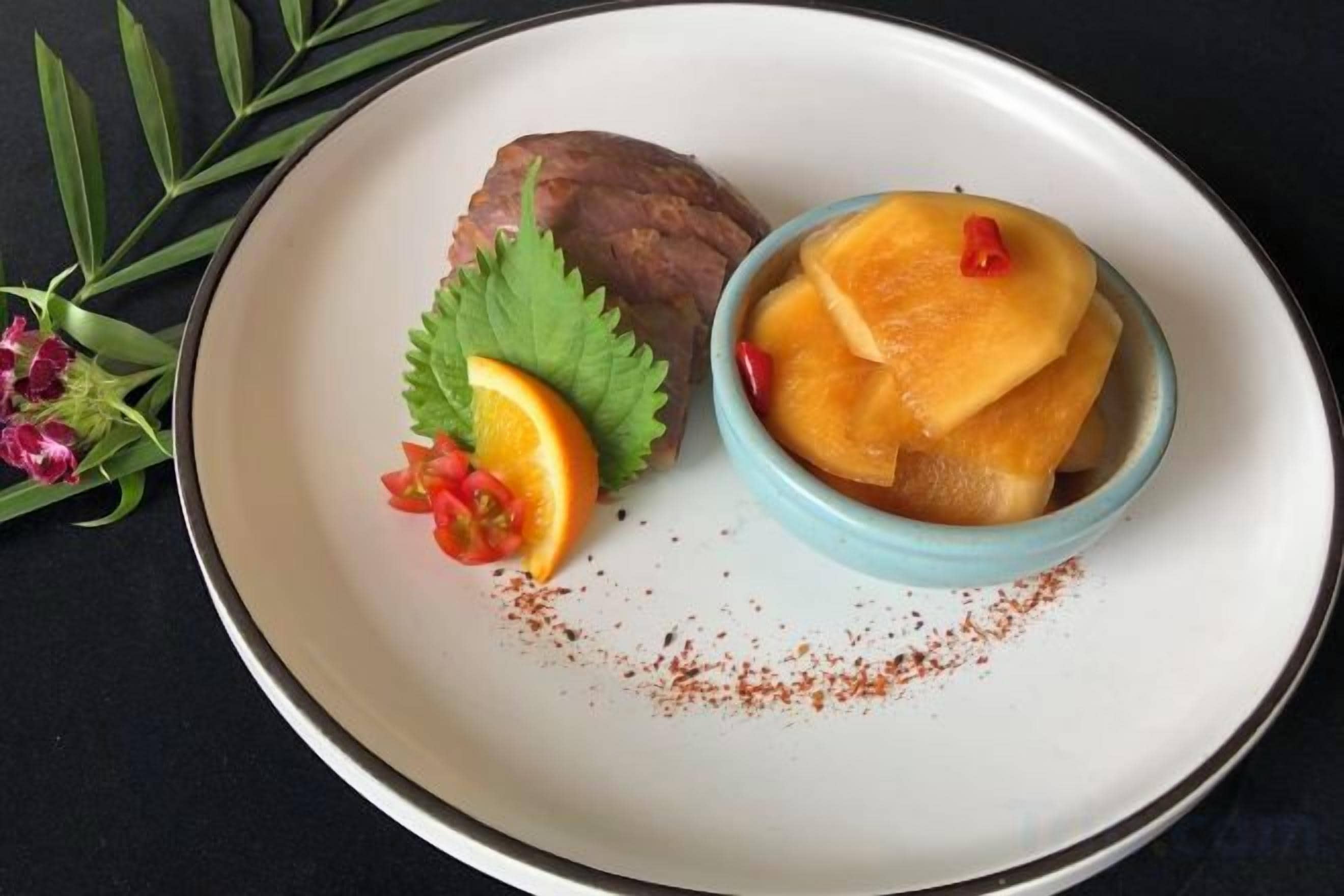 A plate with a bowl of food, a piece of meat, and some garnishes.