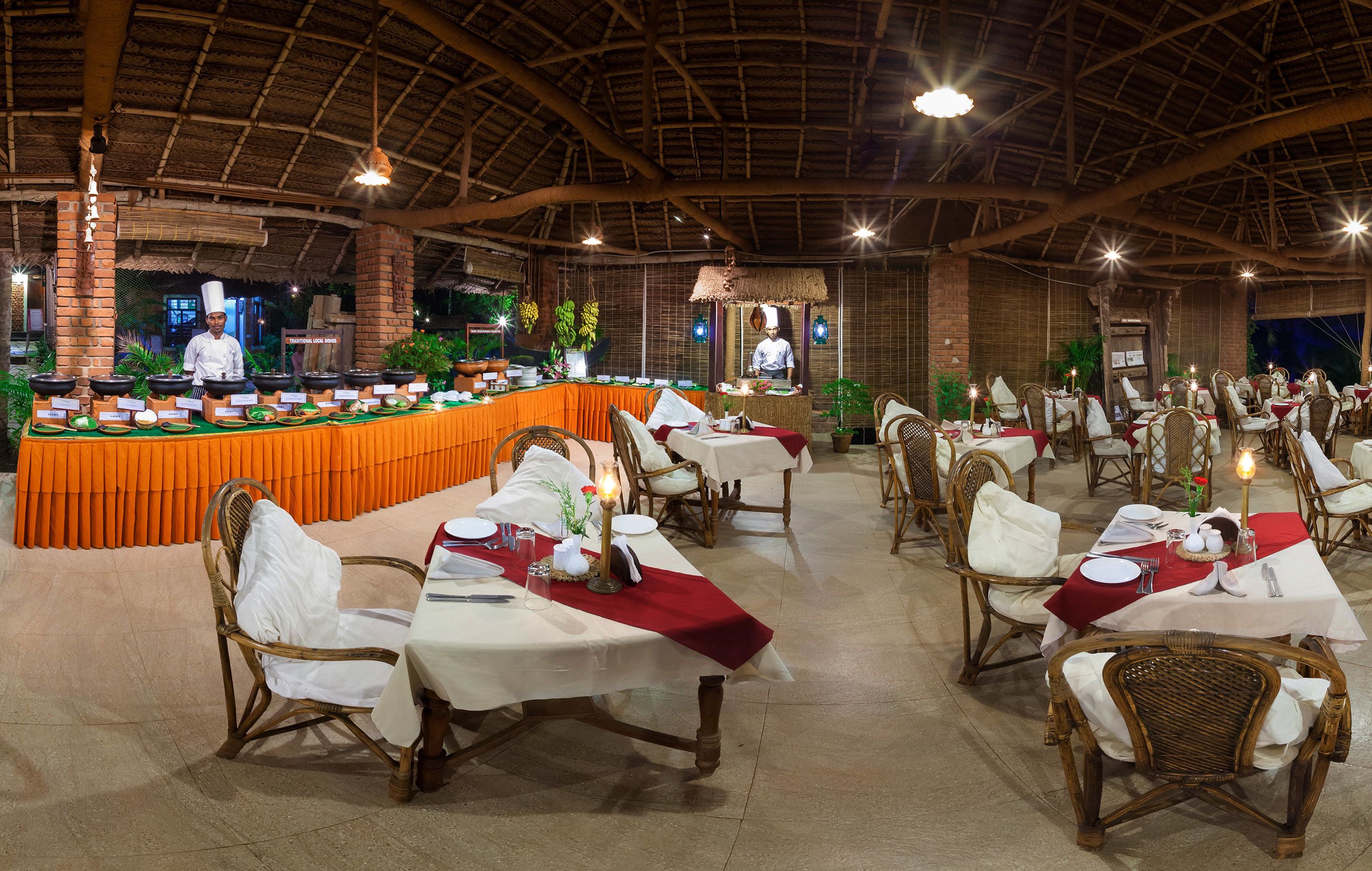 A restaurant with a thatched roof, wicker chairs, and tables set with white tablecloths and red runners.