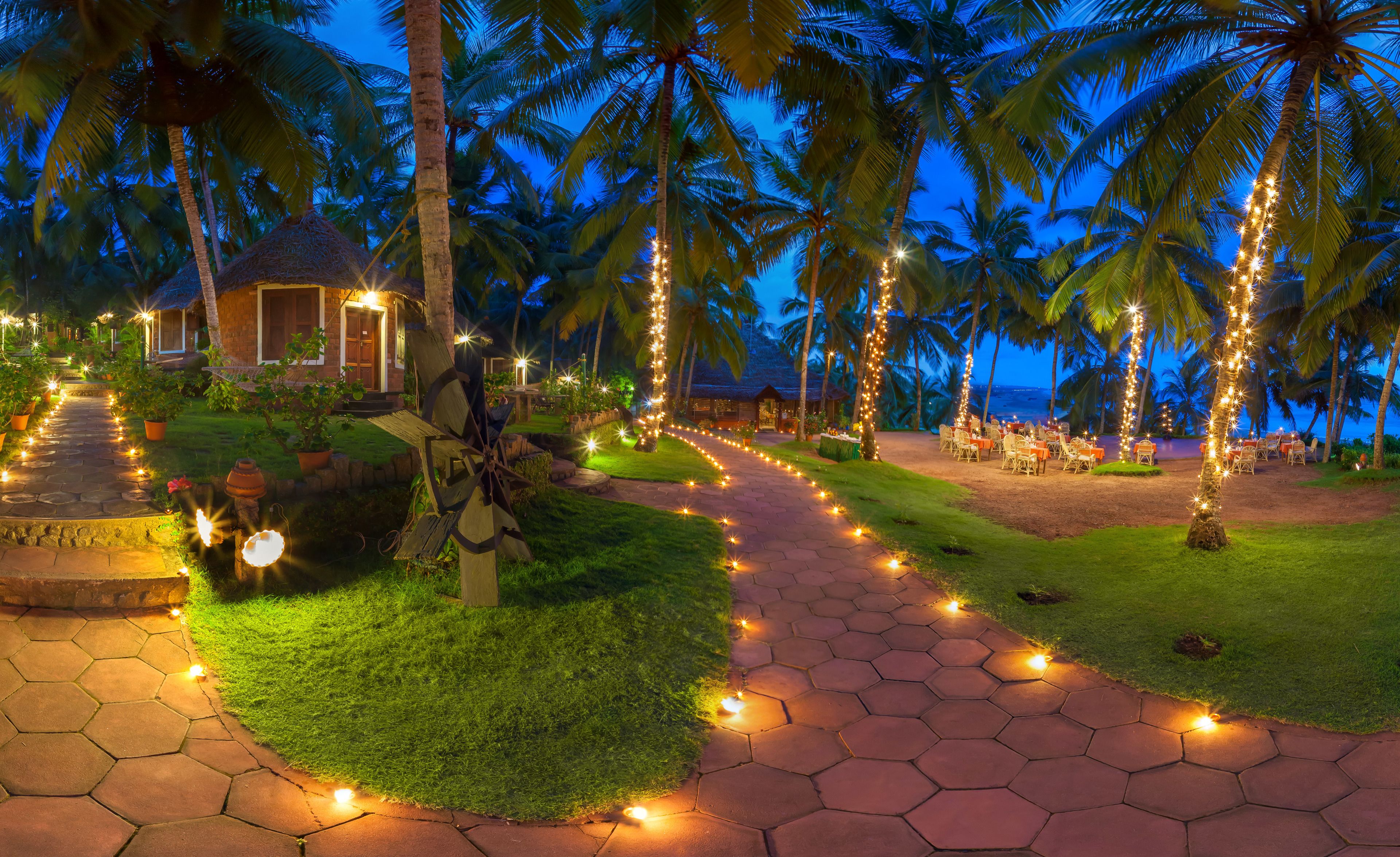 Un resort tropical con un paseo pavimentado bordeado de luces colgantes, palmeras y una zona de playa con mesas y sillas.