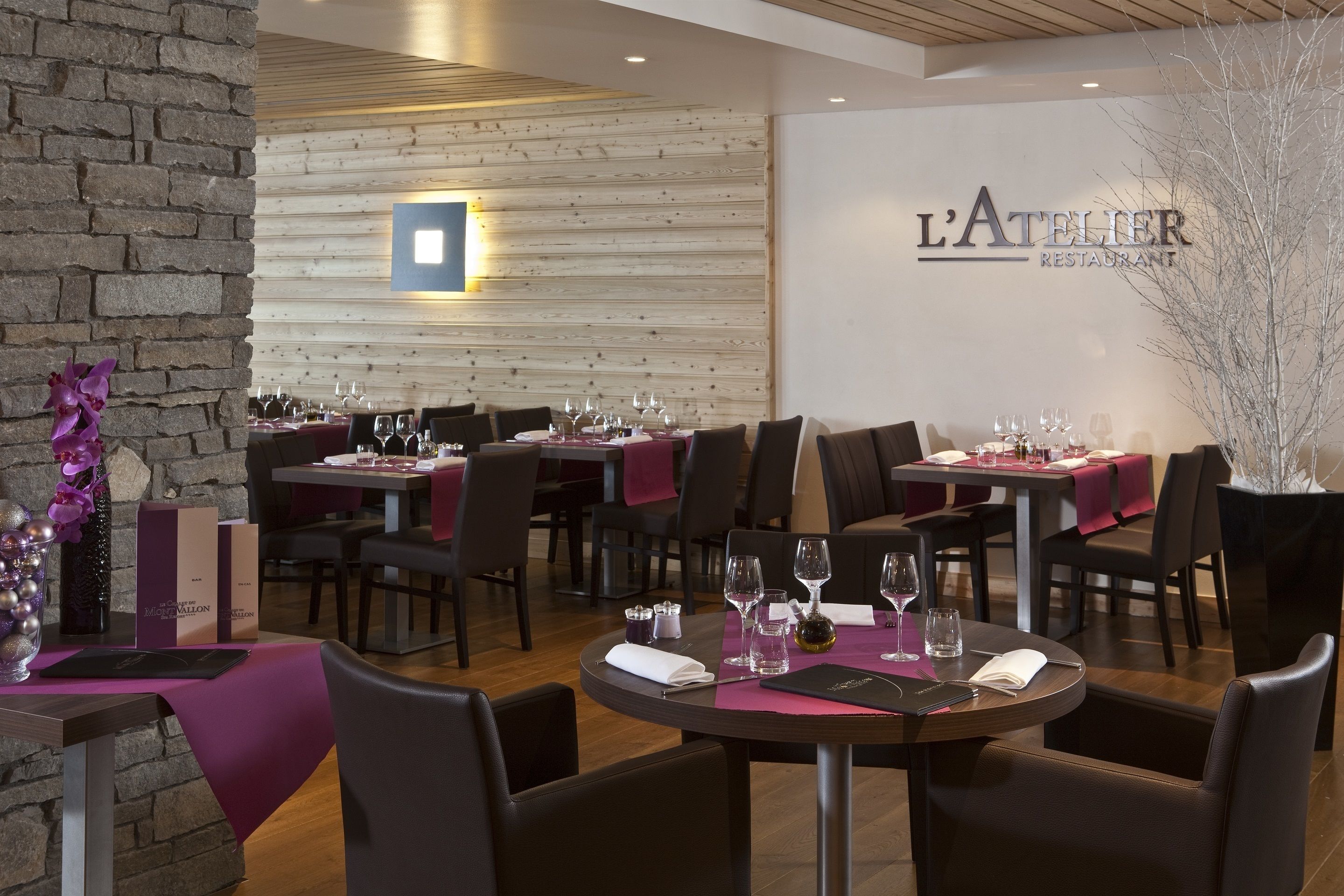 A restaurant interior with wooden panel walls, tables set with wine glasses, and a sign reading "L'Atelier Restaurant".
