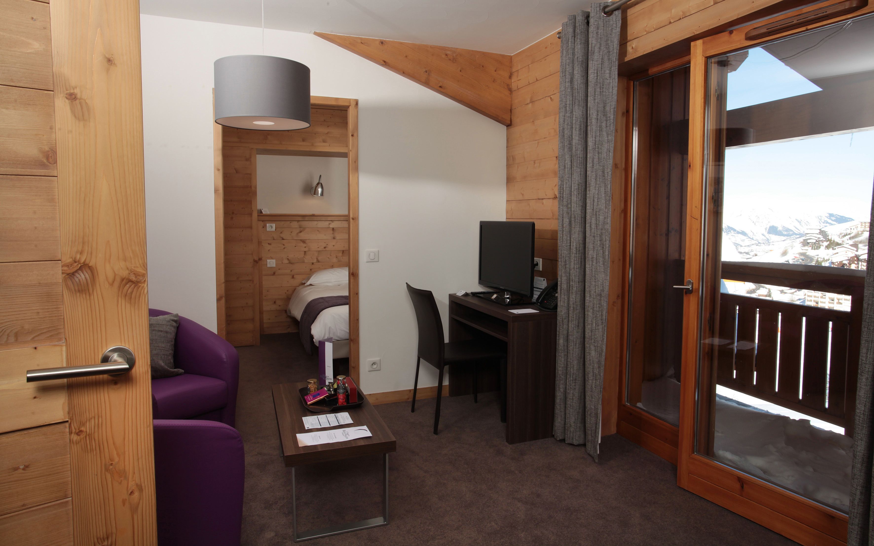 Een kamer met een bed, een bureau met een computer, een stoel, een paarse bank, een klein tafeltje met spullen, en een balkon met uitzicht op bergen.
