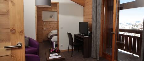 Een kamer met een bed, een bureau met een computer, een stoel, een paarse bank, een klein tafeltje met spullen, en een balkon met uitzicht op bergen.