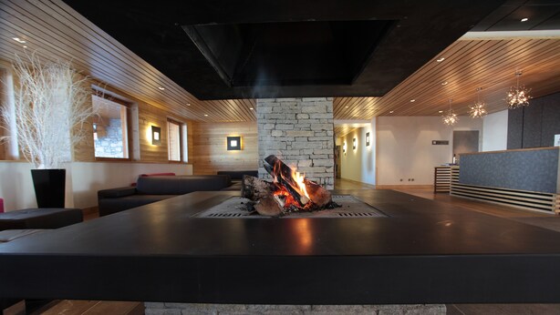 A modern lobby with a central fire feature, wooden paneling, and a stone accent wall. - Residence Le Chalet du Mont Vallon (Les Belleville)