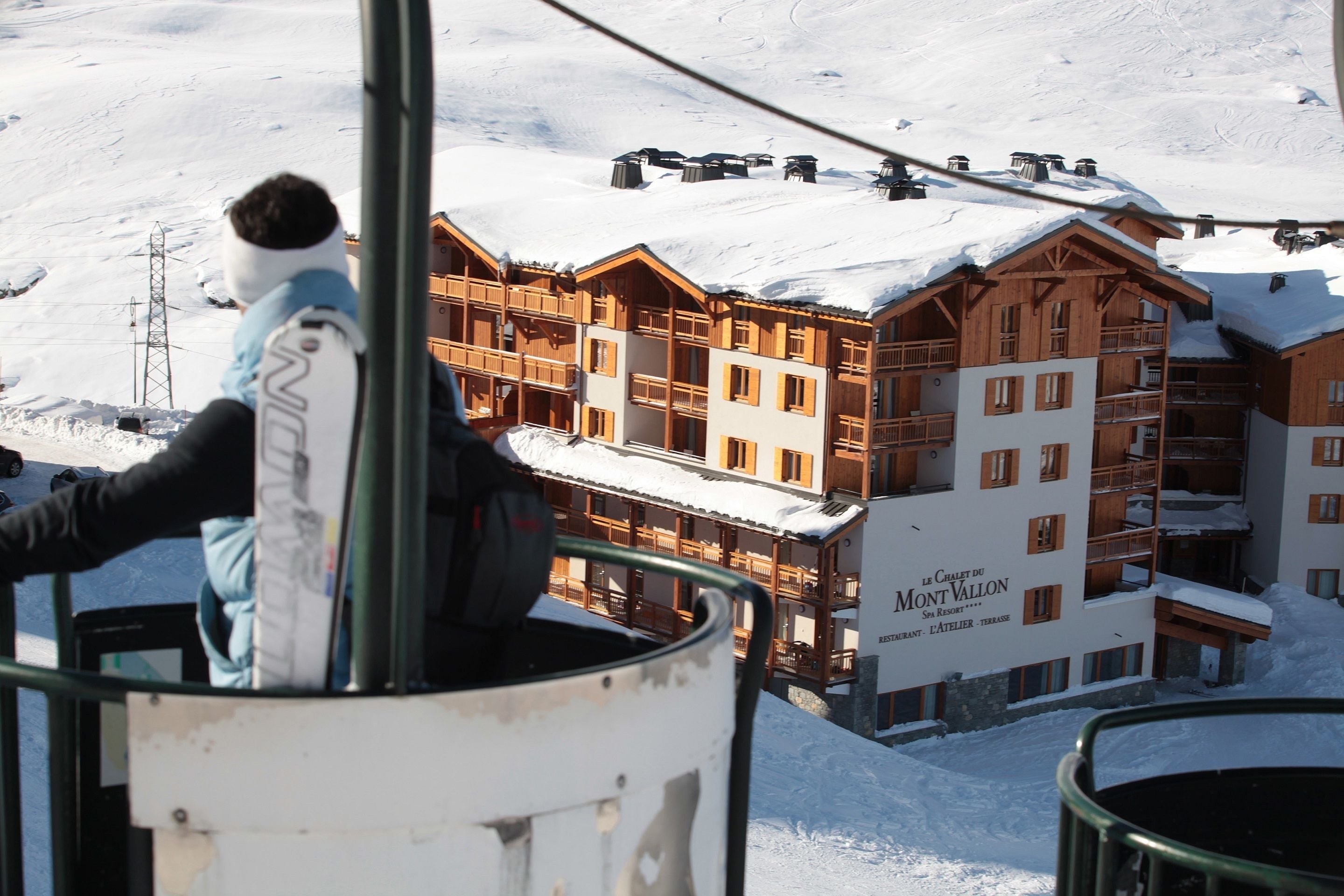 A ski lift chair with a person in a ski jacket. In the background, there is a large wooden building with multiple balconies and a sign that reads "Le Chalet du Mont Vallon."