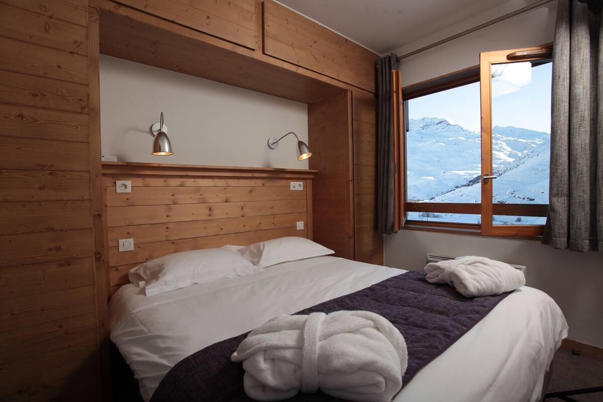 Una habitación con paneles de madera, una cama, dos lámparas de pared y un ventanal con vistas a la montaña.