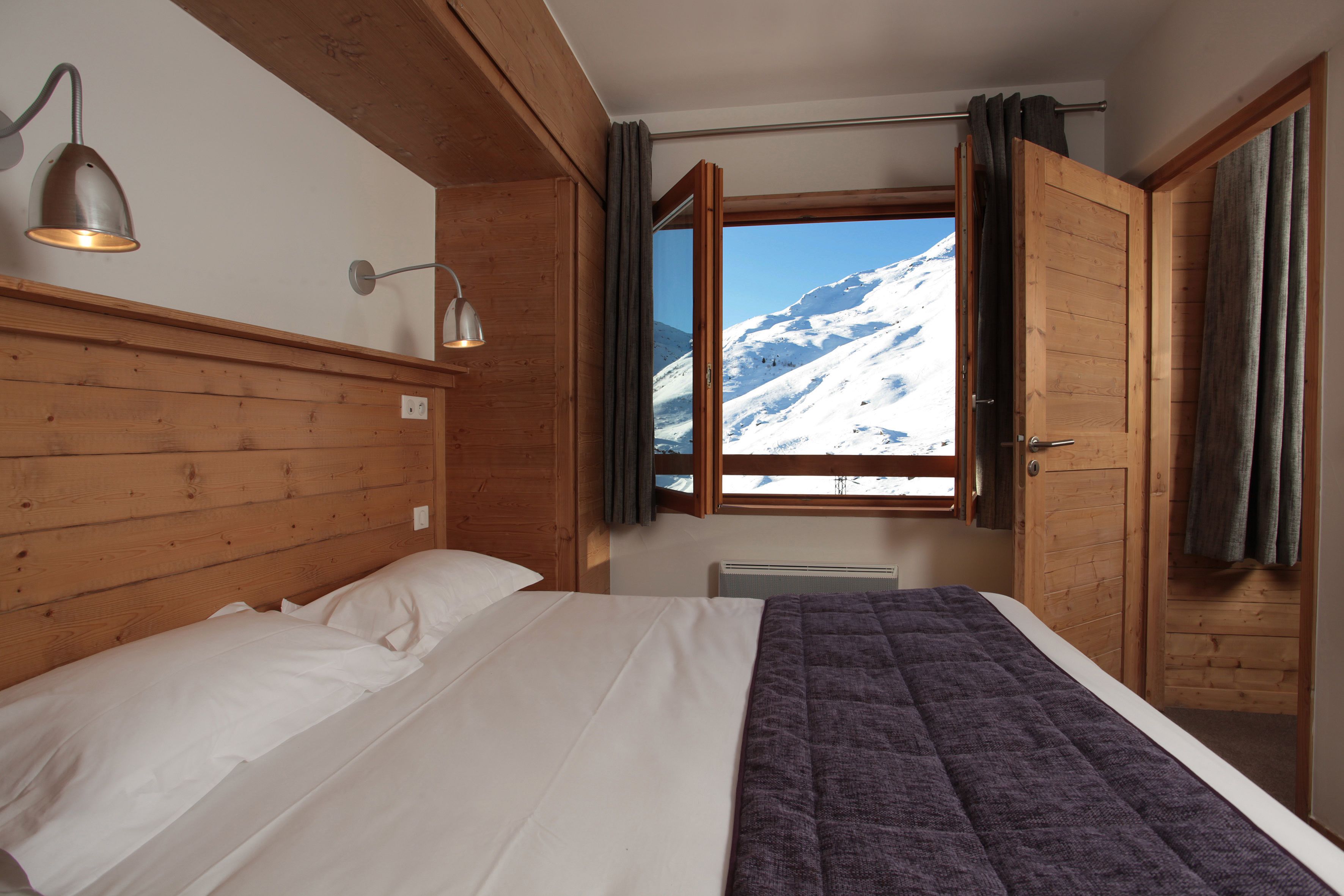 Ein Schlafzimmer mit einem Bett, einem Holzkopfteil, zwei Nachttischlampen, einem Fenster mit Vorhängen und Blick auf einen verschneiten Berg.