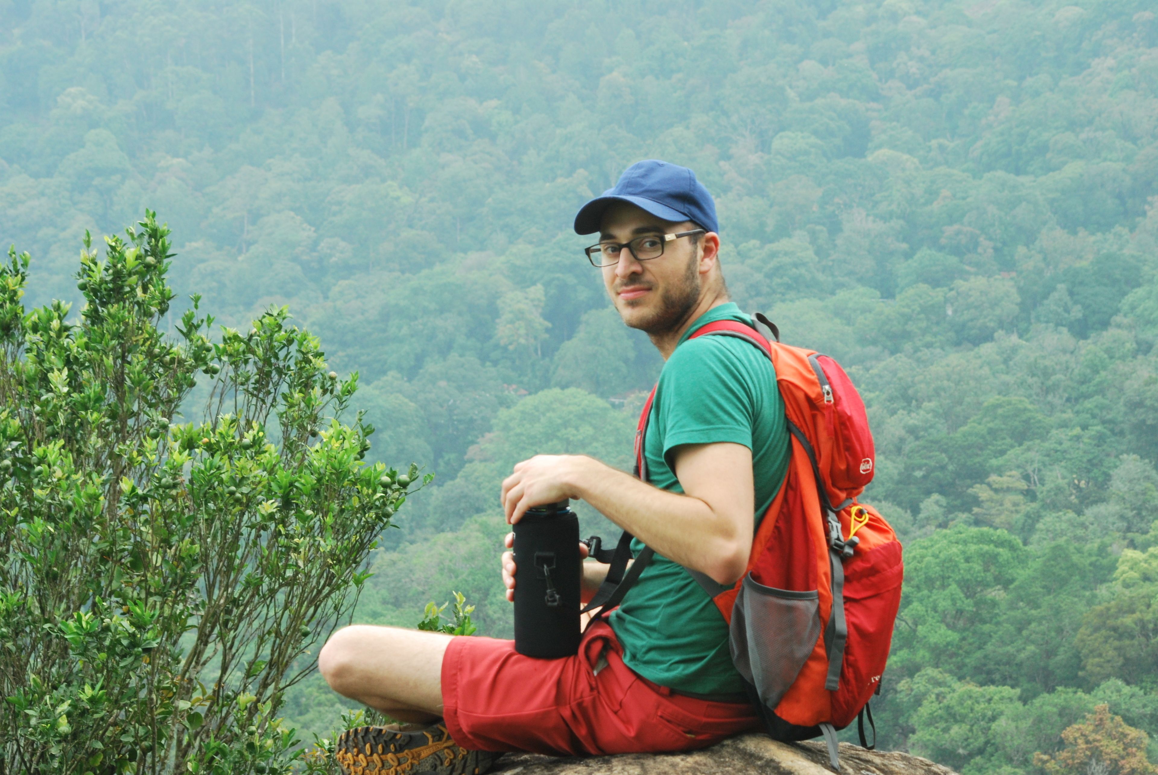 Une personne assise sur un rocher, avec un sac à dos et une bouteille d’eau, entourée d’une forêt dense.