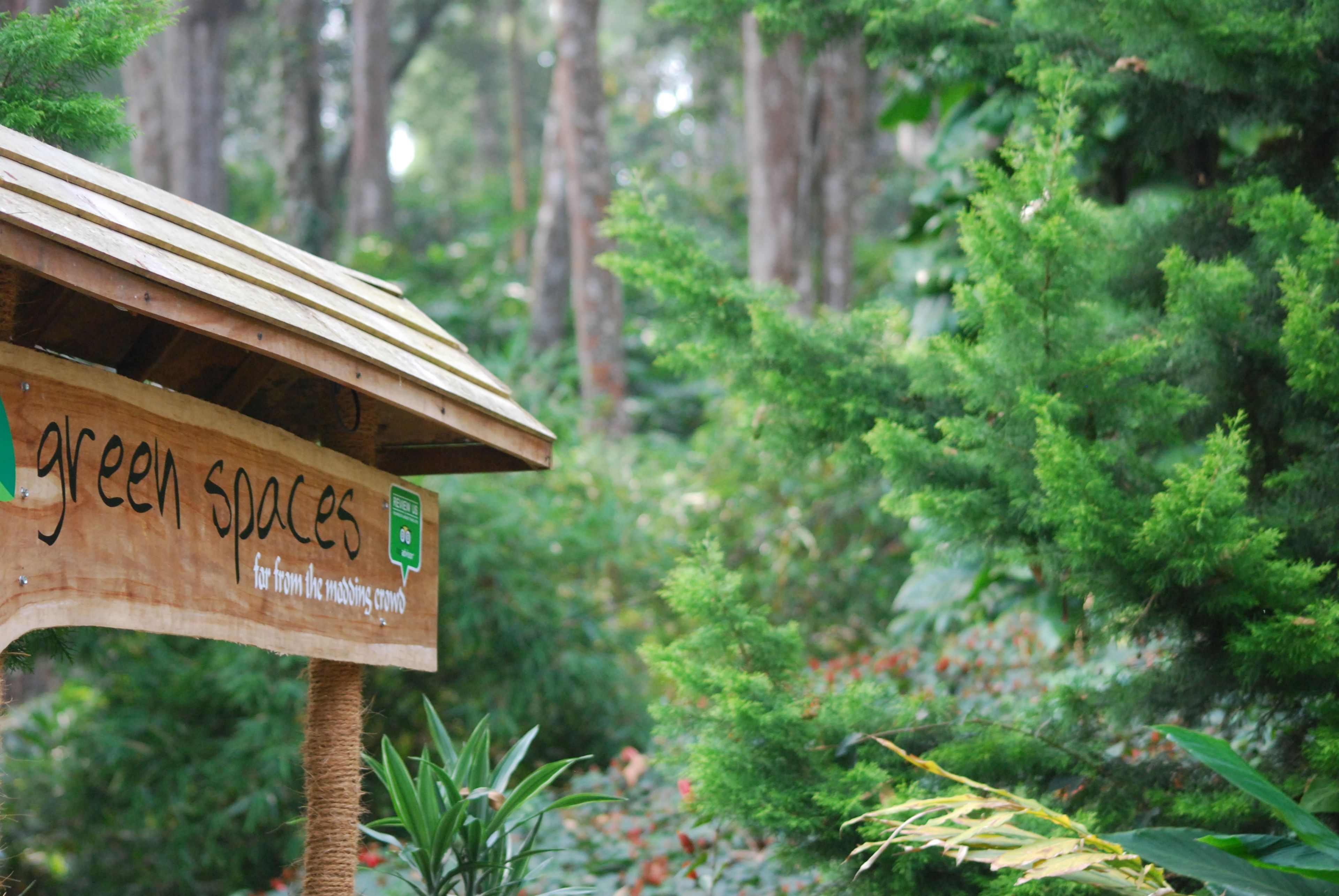 A wooden sign with the text "green spaces" and a logo, set against a backdrop of dense green foliage and trees.