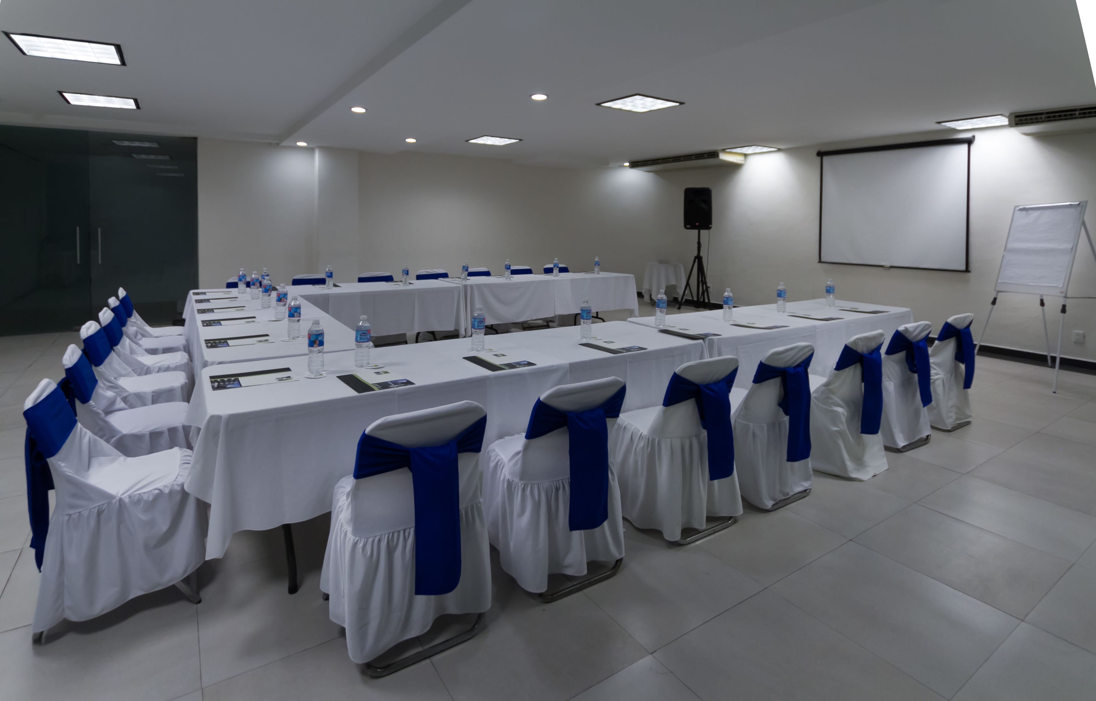 A conference room with long tables, chairs with blue sashes, a projector screen, and a whiteboard.