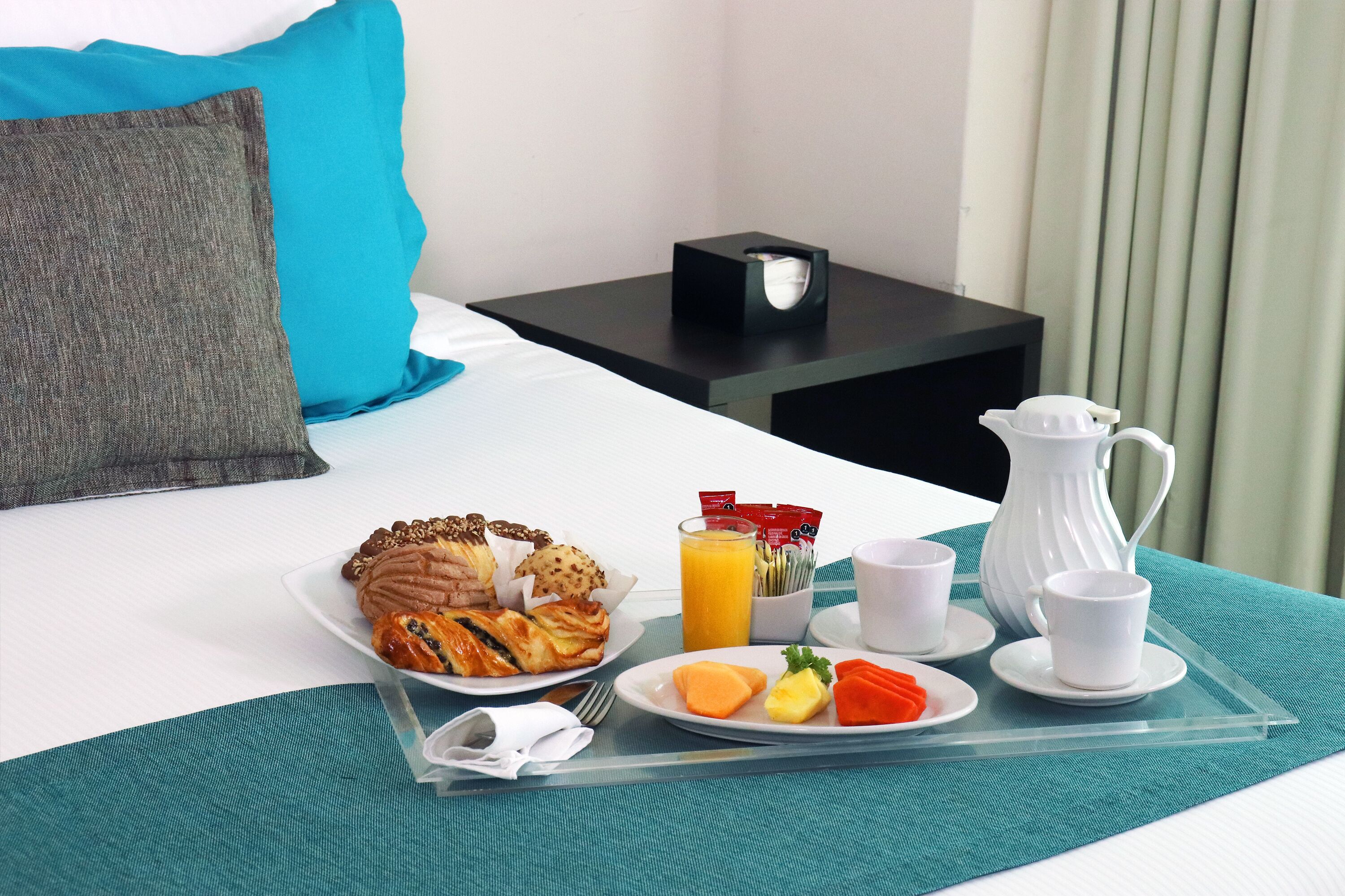 A neatly made bed with a teal bedspread, a breakfast tray with croissants, fruit, and coffee cups, and a white pitcher on a bedside table.