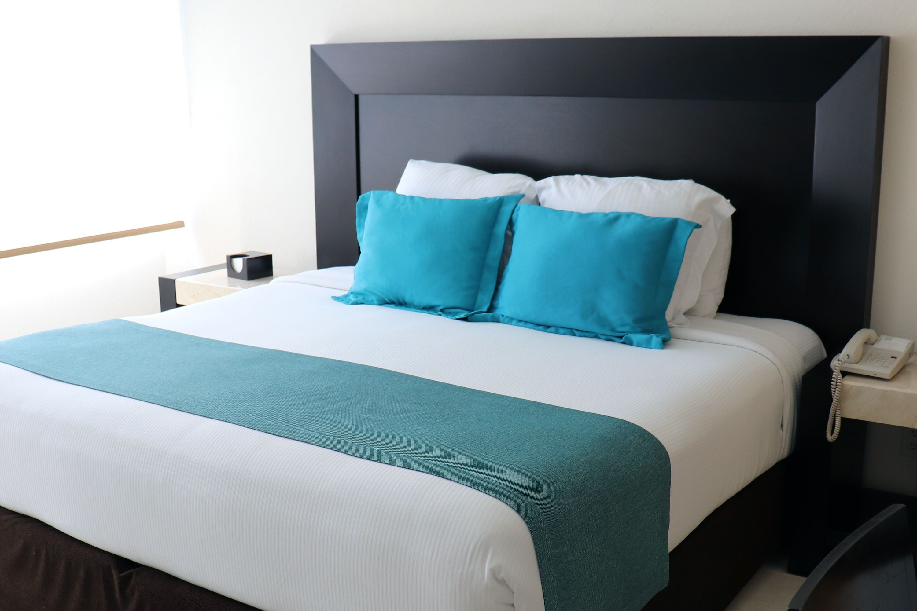 Una cama bien hecha con una colcha azul aguamarina y blanca, un cabecero oscuro y un teléfono en la mesita de noche.