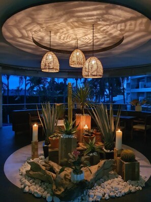 Un espacio interior bien iluminado con una zona de estar circular rodeada de plantas en macetas y velas, con un techo de diseño texturizado único.