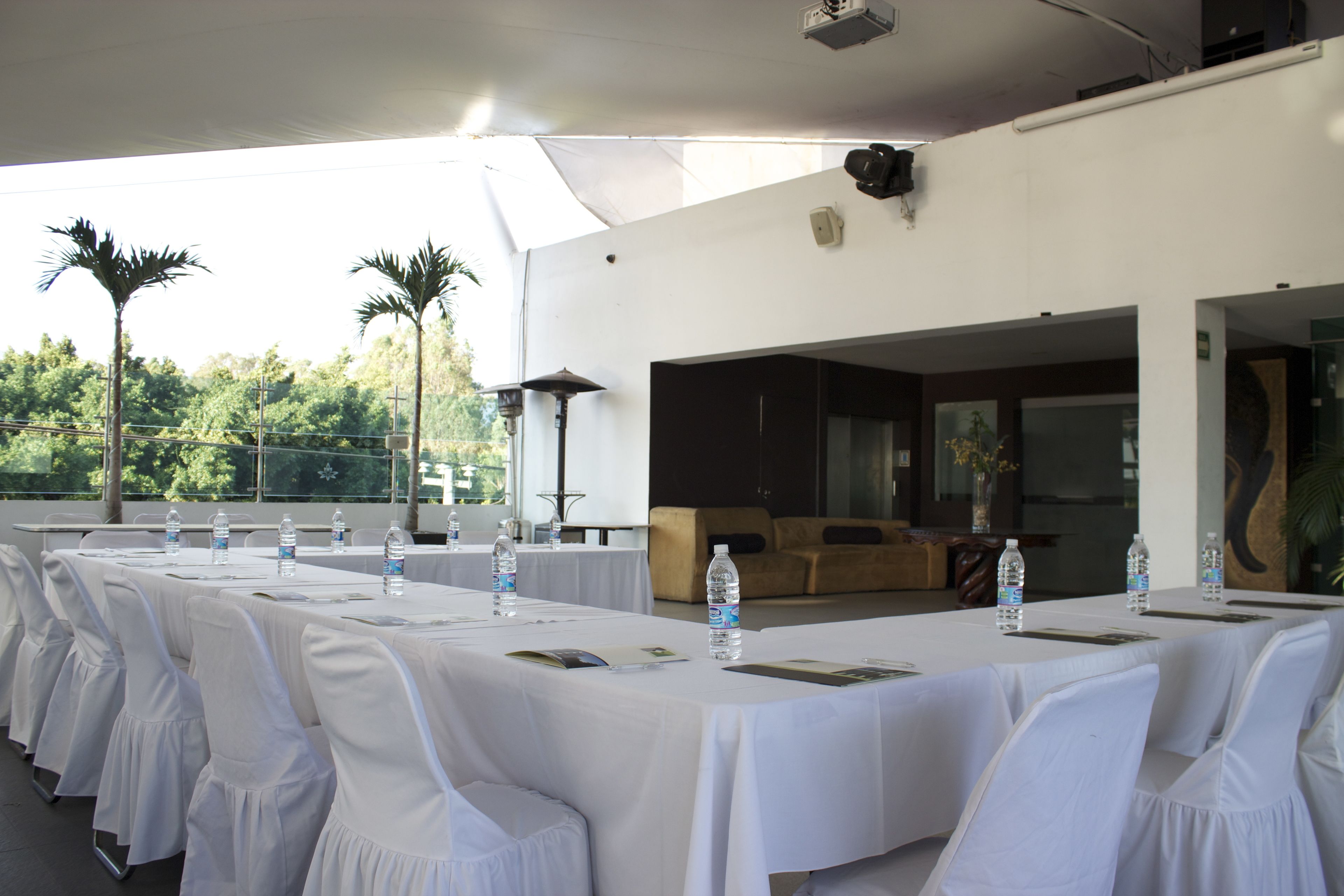 A covered outdoor dining area with white tablecloths and chairs, water bottles, and a view of trees and a building in the background.