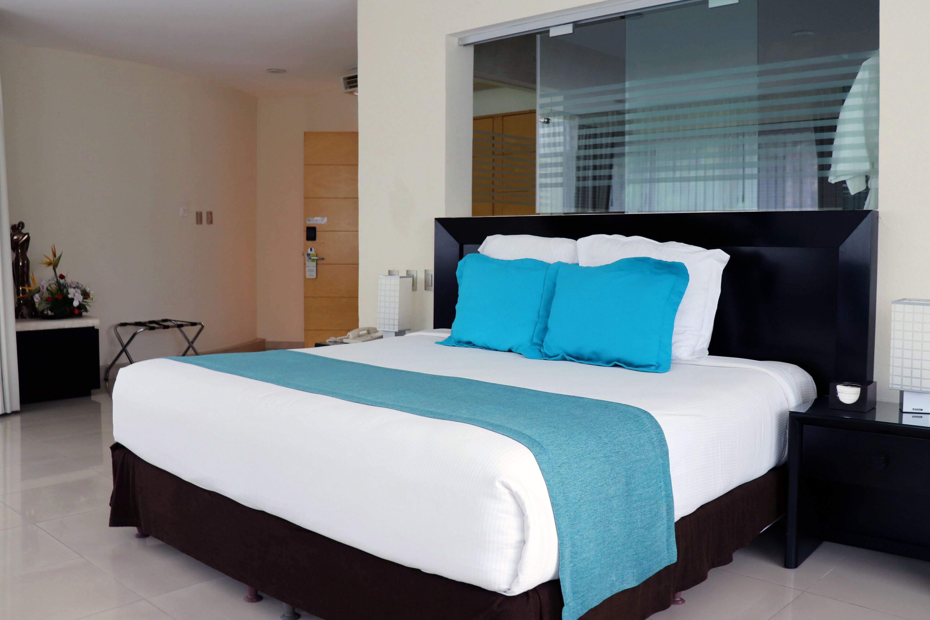 A modern hotel room with a large bed, a nightstand, a mirror, and a view of the outside through the window.