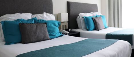 Two twin beds with teal and gray bedding, a bedside table with a lamp, and a window with curtains.