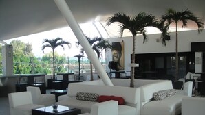A modern lounge area with white sofas, ottomans, and a black coffee table. There are palm trees and a large painting on the wall. - Hotel Río 1300 (Cuernavaca)