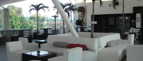 A modern lounge area with white sofas, ottomans, and a black coffee table. There are palm trees and a large painting on the wall.