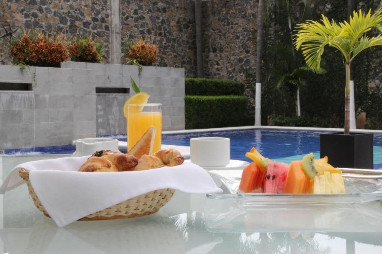 Desayuno con croissants, fruta y jugo de naranja junto a una piscina, con un muro de piedra y vegetación al fondo.