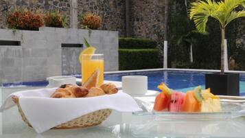 Desayuno con croissants, fruta y jugo de naranja junto a una piscina, con un muro de piedra y vegetación al fondo.