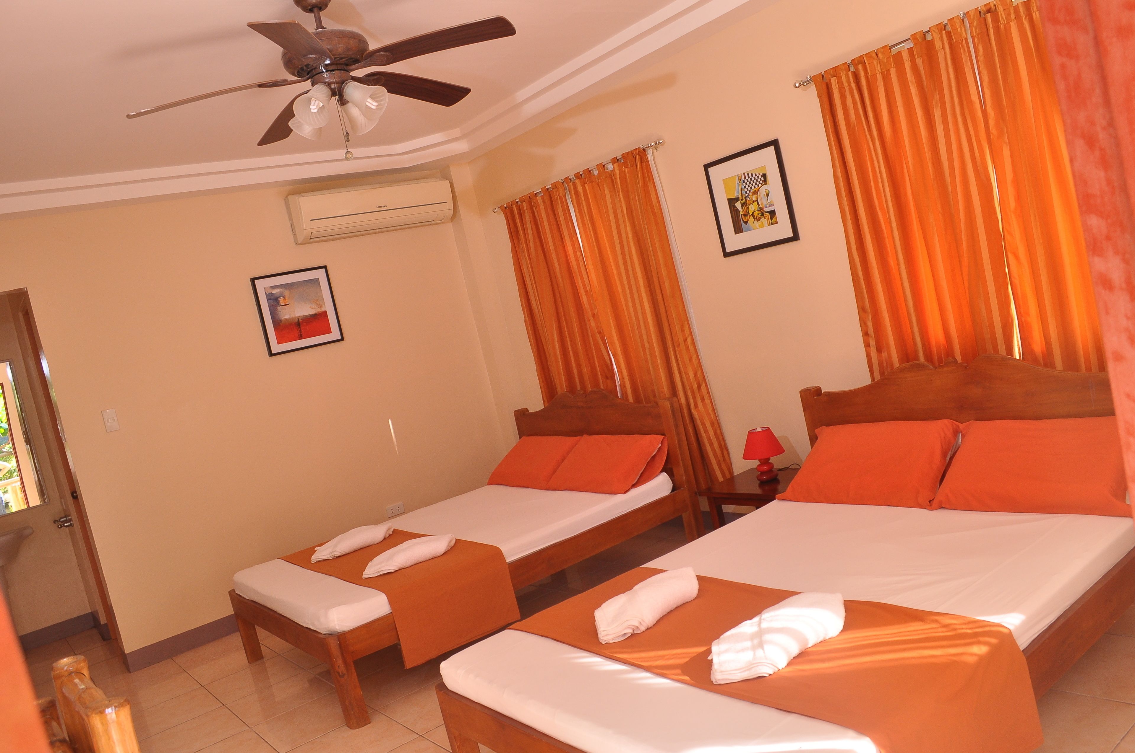 A hotel room with two single beds, a ceiling fan, an air conditioning unit, and orange curtains.