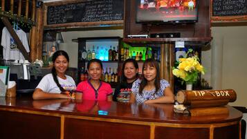 Un bar con mostrador de madera, un televisor montado encima y una pizarra con precios de bebidas.