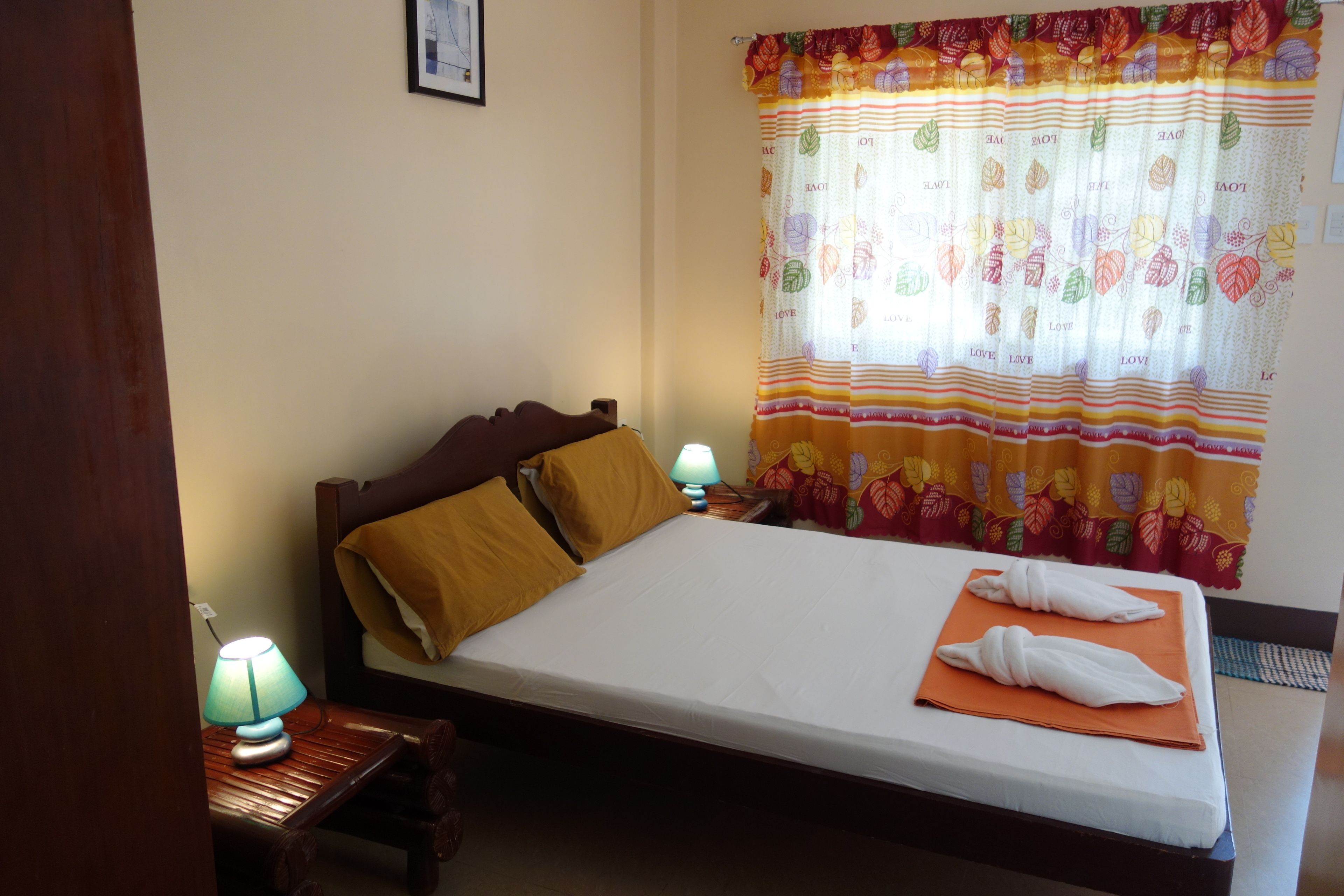 A neatly made bed with a white mattress, two pillows, and a folded towel on an orange cloth. A wooden nightstand with a blue lamp is beside the bed.