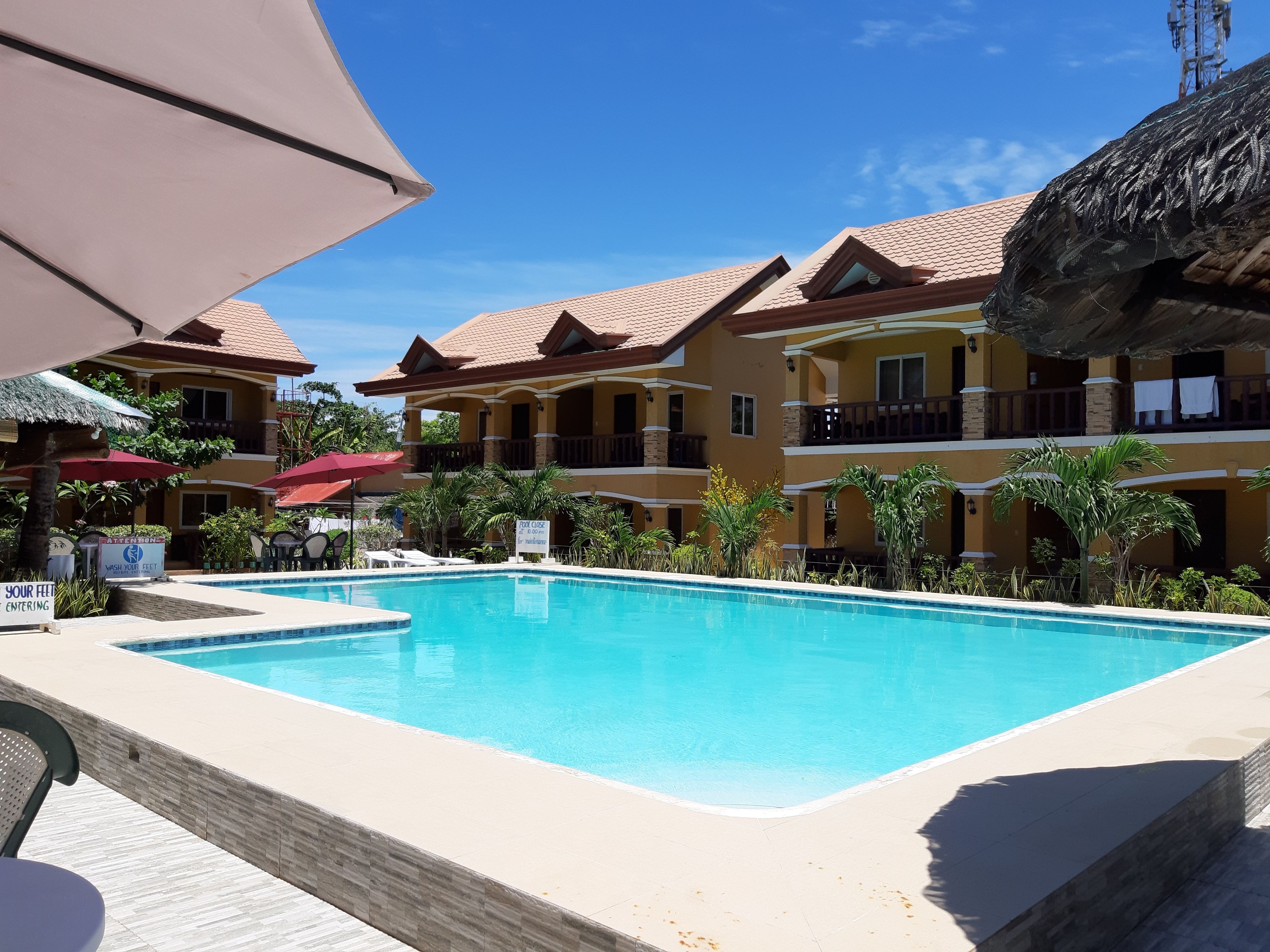 A swimming pool surrounded by multi-story buildings with balconies, palm trees, and a clear blue sky.