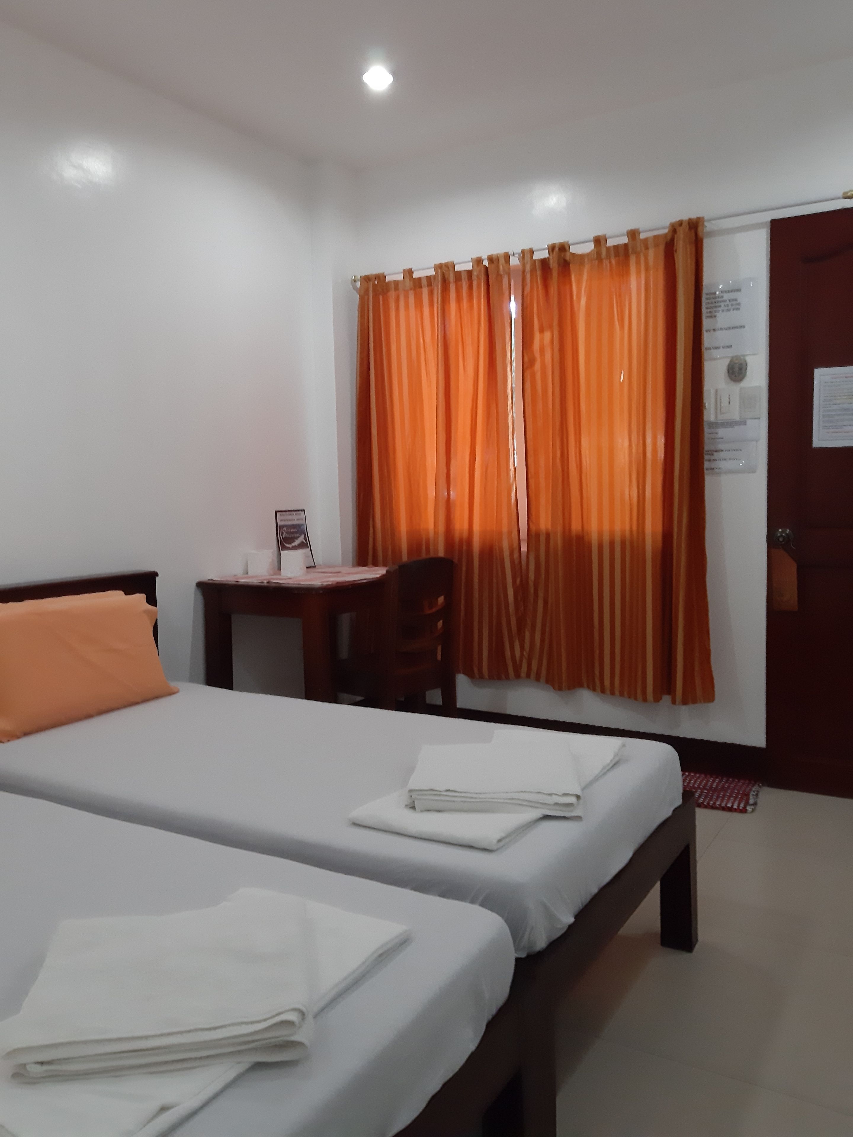 A hotel room with two beds, a desk, a chair, and a window with orange curtains.