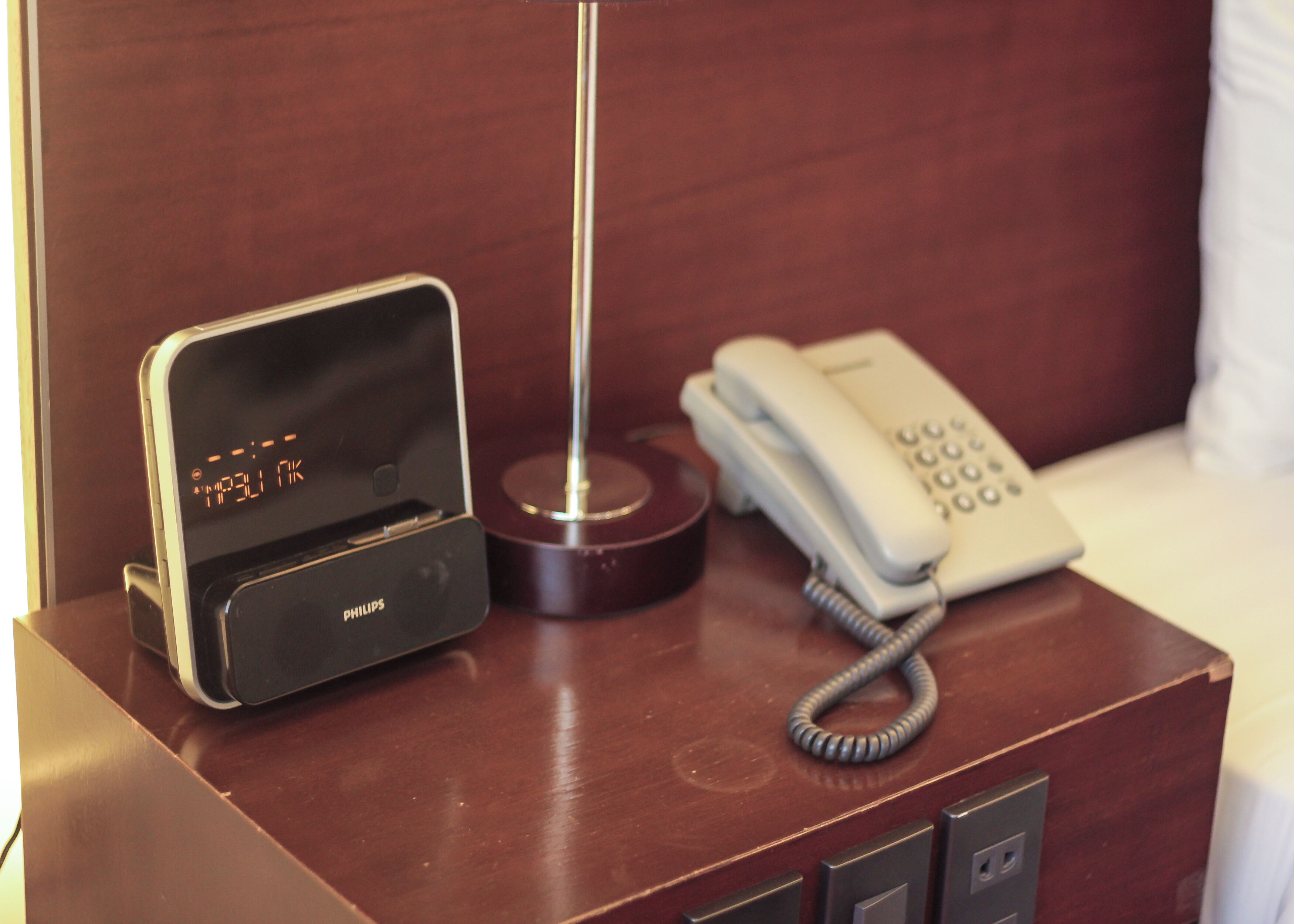Una mesita de noche de madera con un reloj digital Philips, un teléfono y una lámpara.