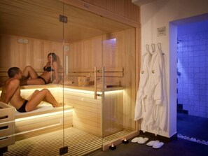 A sauna room with wooden benches and a glass partition. There are towels and slippers on the floor.