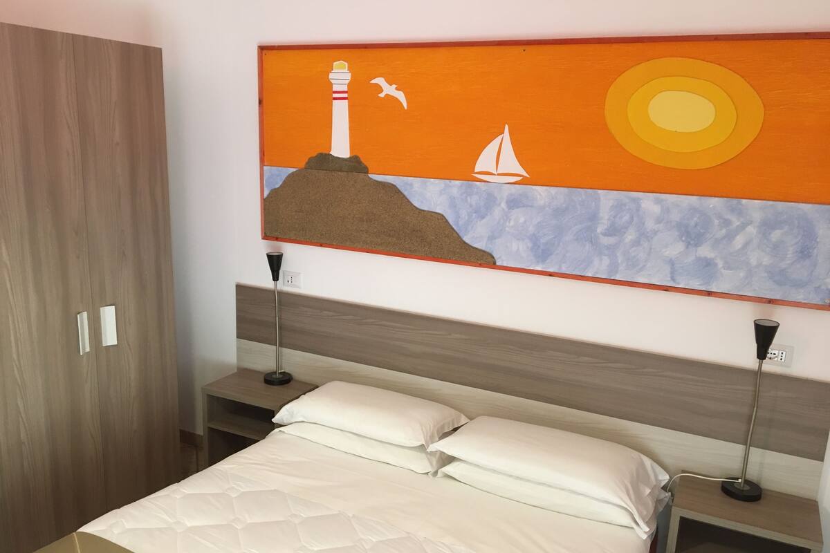 Un dormitorio con cama, dos mesitas de noche, un armario y una obra de arte en la pared que representa un faro, un velero y una gaviota.