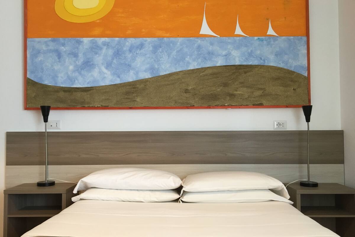 Una habitación de hotel con una cama, dos mesitas de noche y un cuadro colgado en la pared que representa un atardecer sobre un paisaje.