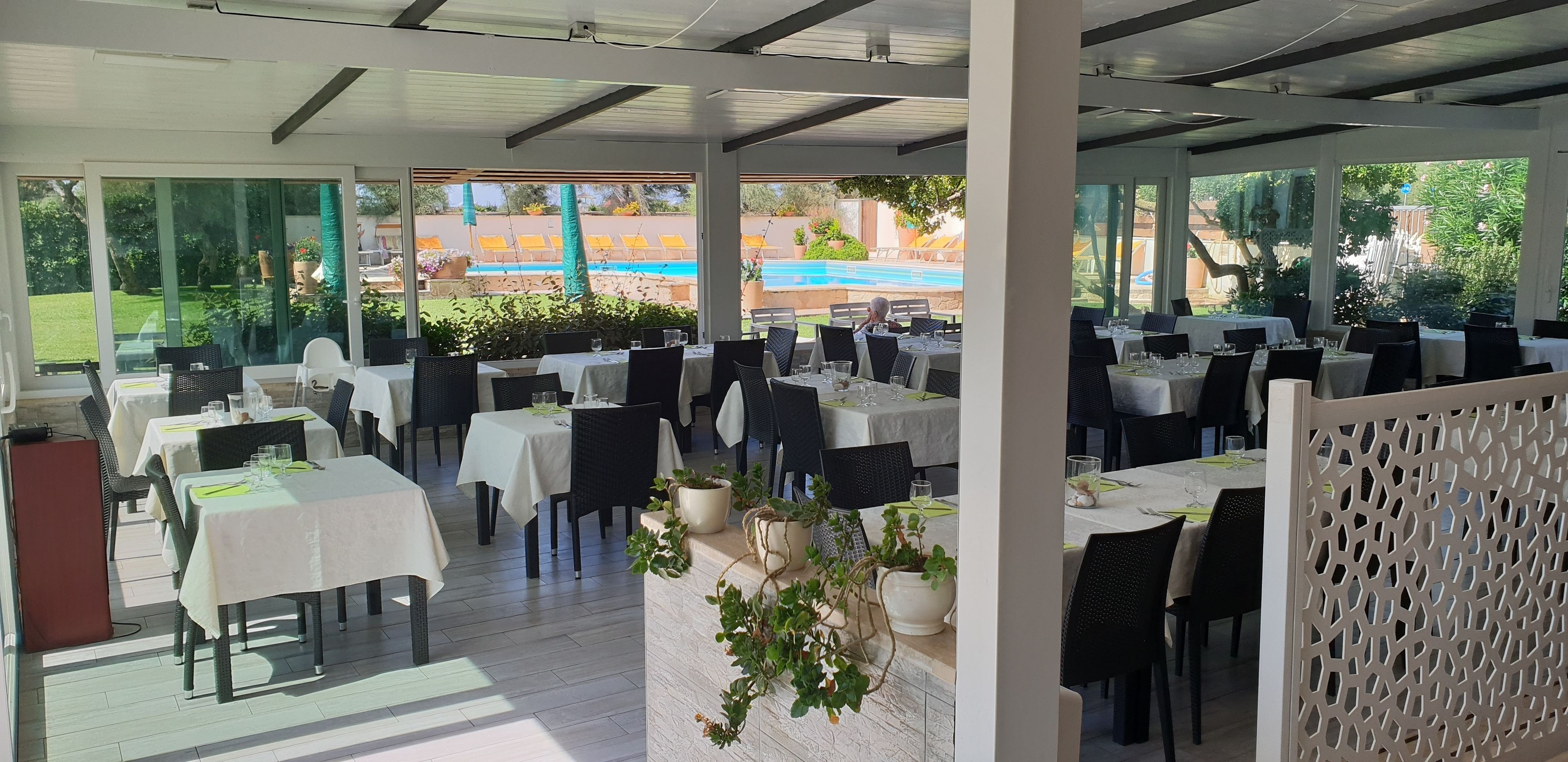 An outdoor dining area with tables set for two, surrounded by potted plants and a pool view.