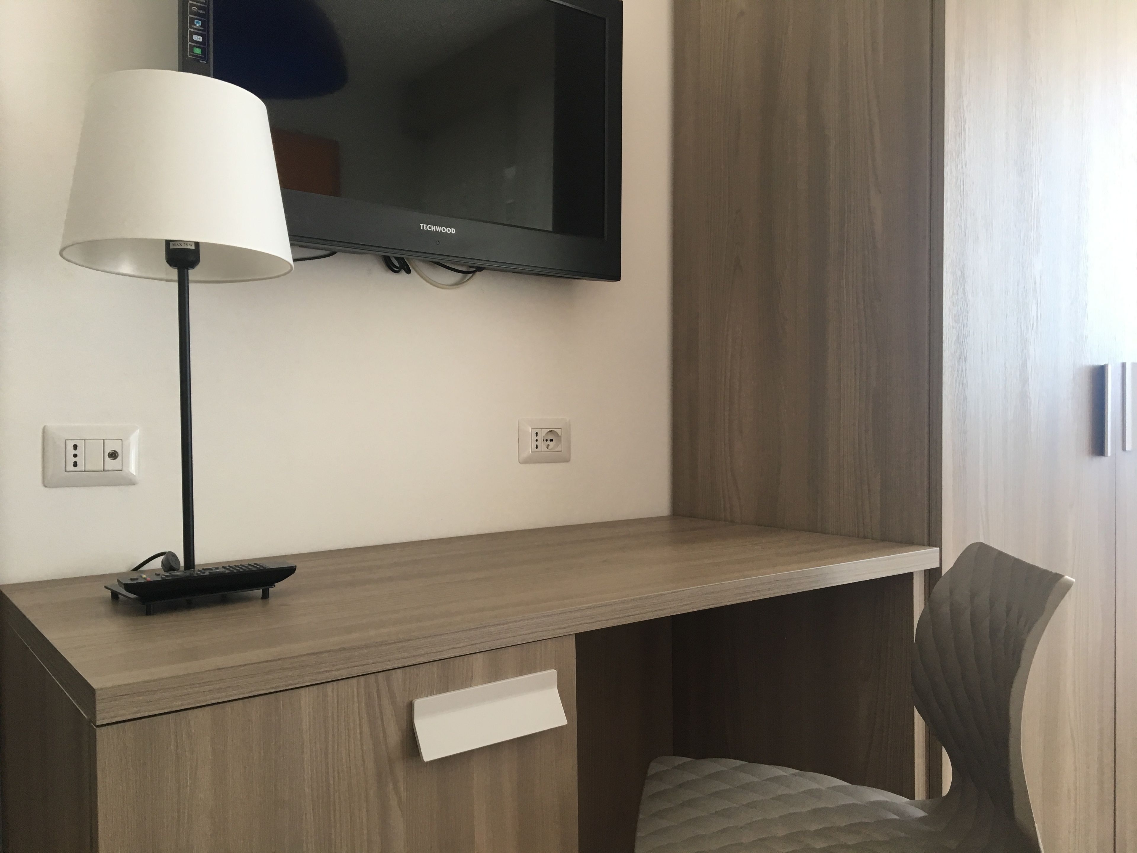 Camera d'albergo con televisore a schermo piatto fissato al muro, una scrivania con lampada, un telecomando e una sedia.