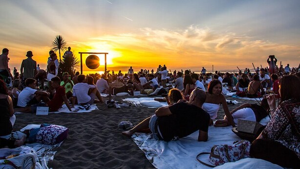 Eine Strandansicht mit Menschen, die auf Handtüchern unter einer großen Sonnendachkonstruktion sitzen, vermutlich für eine Sonnenveranstaltung.