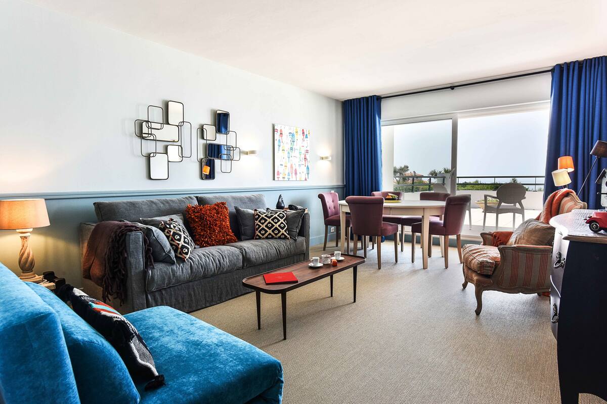 Una habitación de hotel moderna con sofá gris, sillón azul, zona de comedor con mesa y sillas, y un ventanal con cortinas.