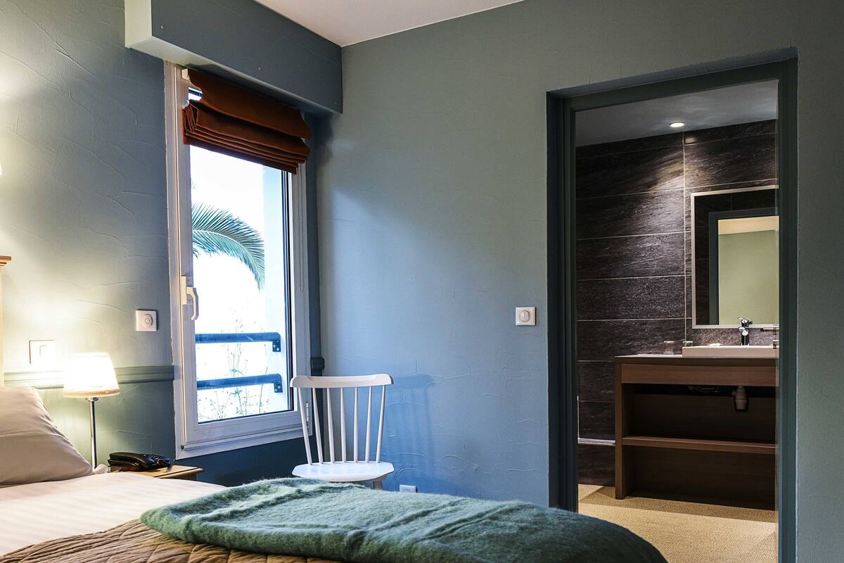 Una habitación de hotel con una cama, una ventana con vistas, un baño con espejo y una revista sobre la cama.