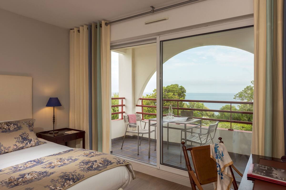 Un dormitorio con cama, mesita de noche, lámpara y vistas a un balcón con mesa y sillas.