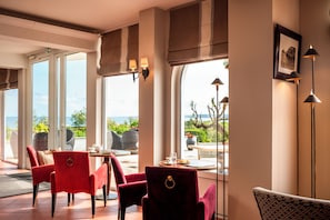 Habitación de hotel con amplios ventanales, una zona de comedor con sillas rojas y vista al mar.