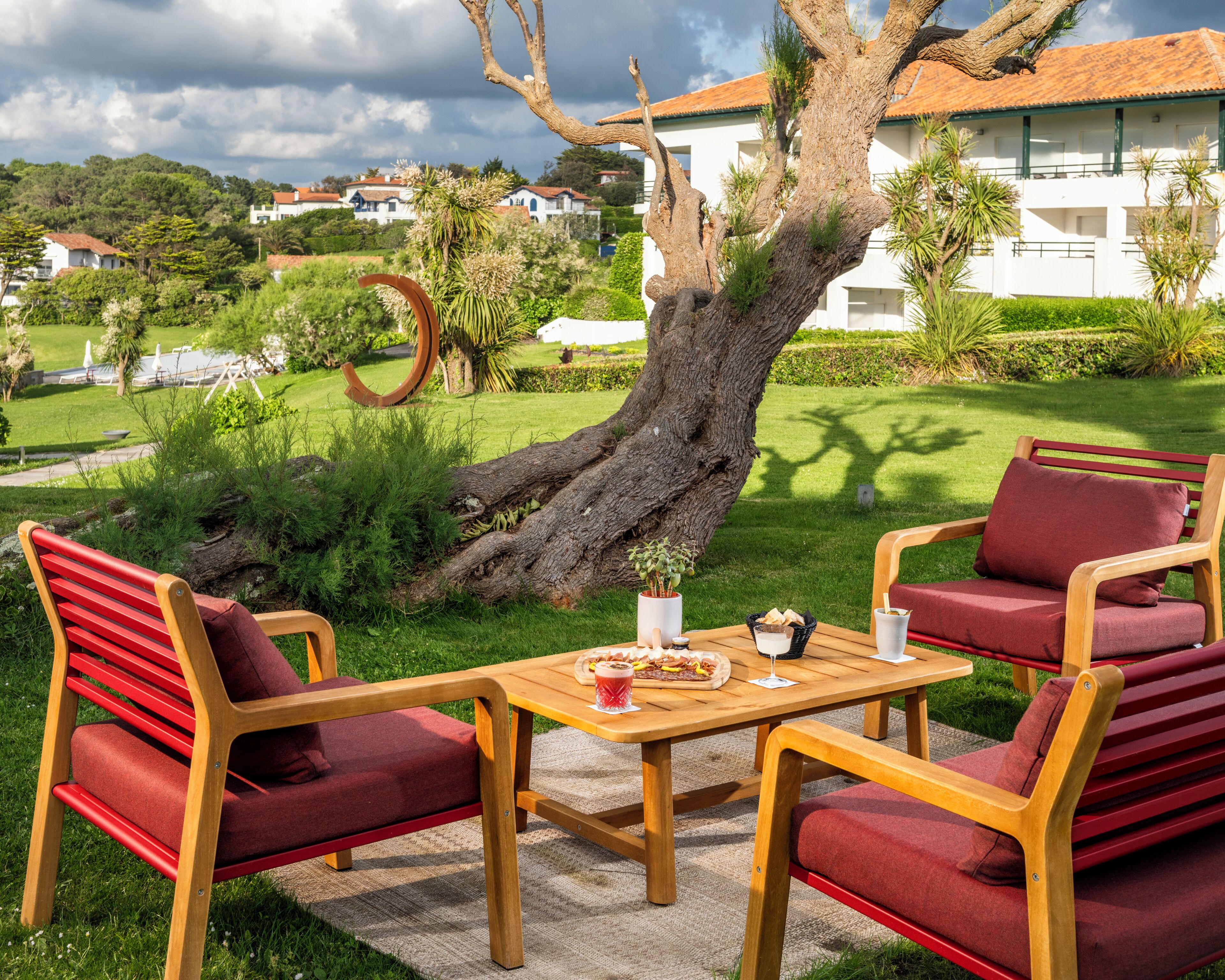 Una mesa de madera con sillas tapizadas rojas, una planta en maceta y un vaso de bebida en un área verde con un árbol grande y un edificio blanco al fondo.