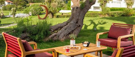 Una mesa de madera con sillas tapizadas rojas, una planta en maceta y un vaso de bebida en un área verde con un árbol grande y un edificio blanco al fondo.