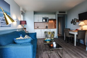 A modern living room with a blue sofa, a coffee table, and a dining area with a table and chairs. A kitchen is visible in the background with white cabinets and a microwave. - Hôtel et Résidence La Réserve (Saint-Jean-de-Luz)