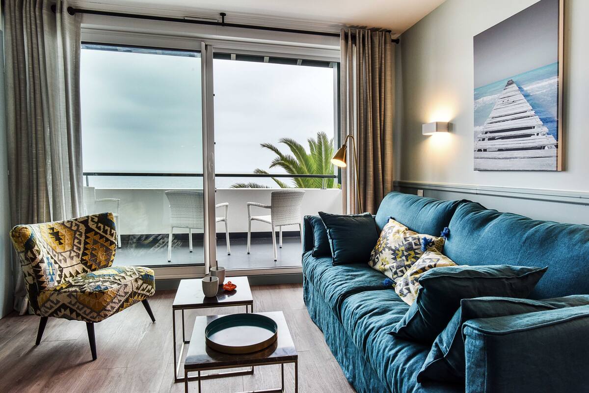 Un salón moderno con un sofá, un sillón y una mesa con un cuenco decorativo. Se ve un balcón con vistas a palmeras a través de la ventana.